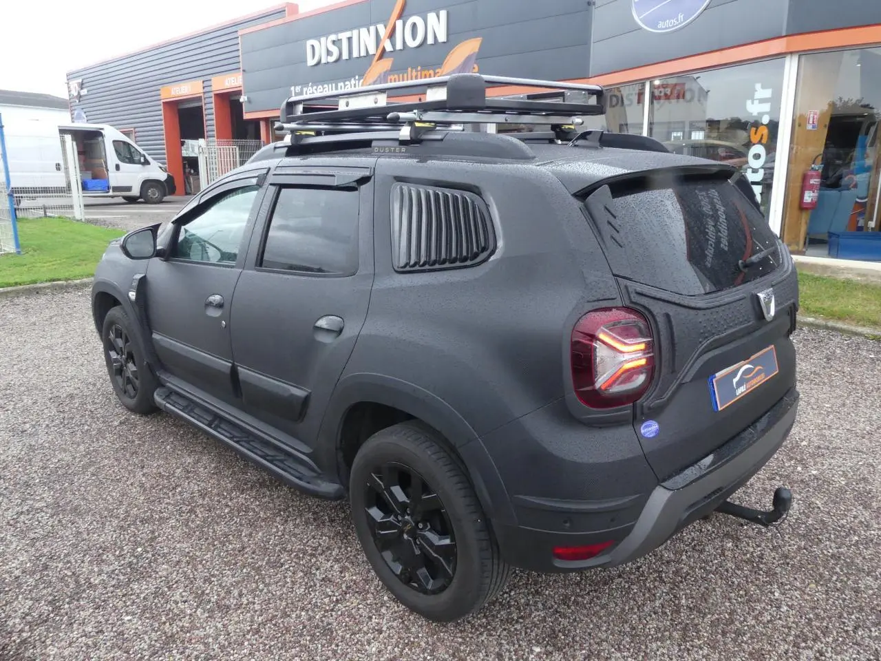 Vue 3/4 arrière droite du Dacia Duster noir 2023 avec covering intégral, galerie de toit et attelage fixe.