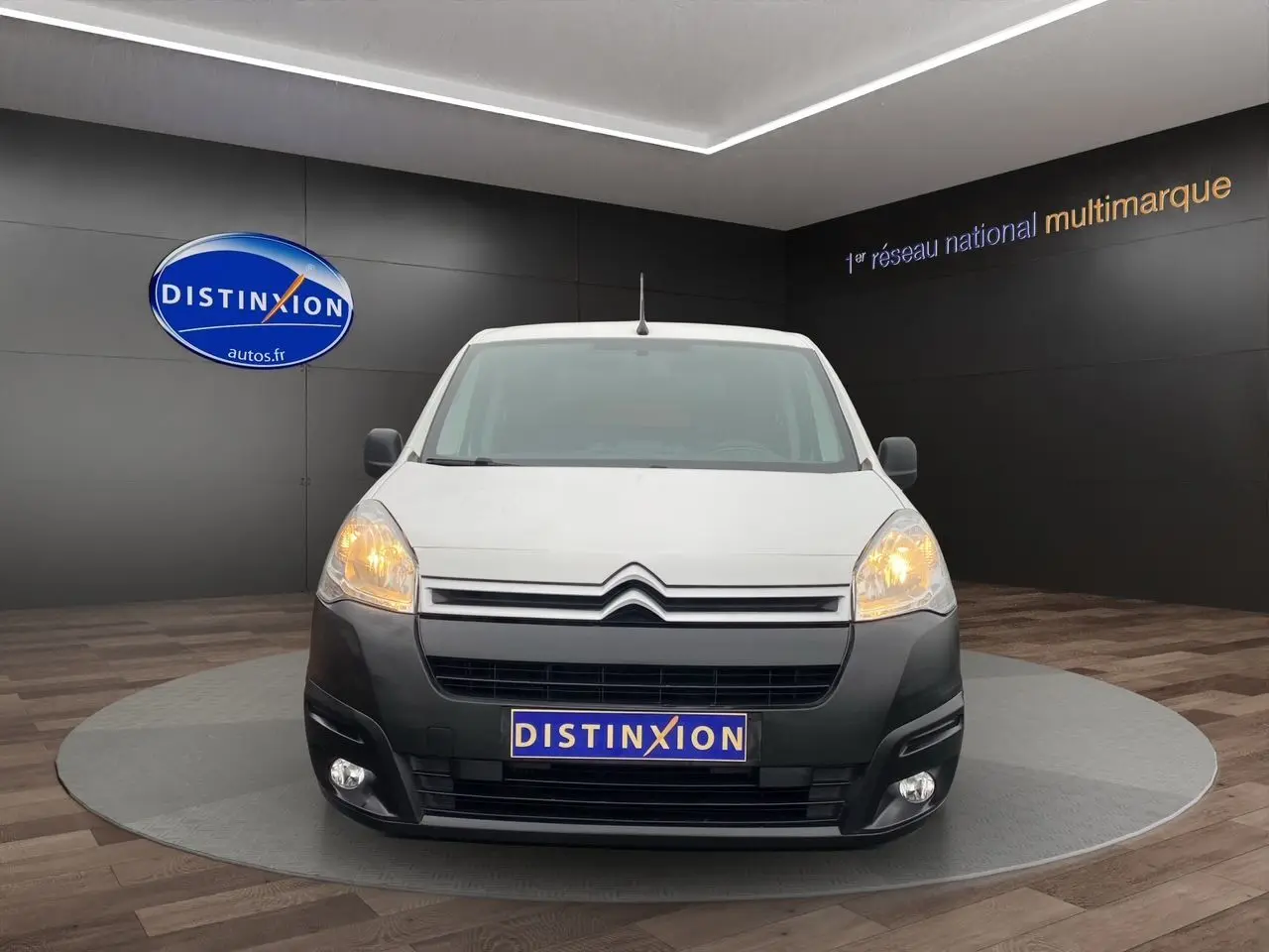 Vue avant d'un Citroën Berlingo blanc 2018 avec pare-chocs noir et phares allumés en showroom.