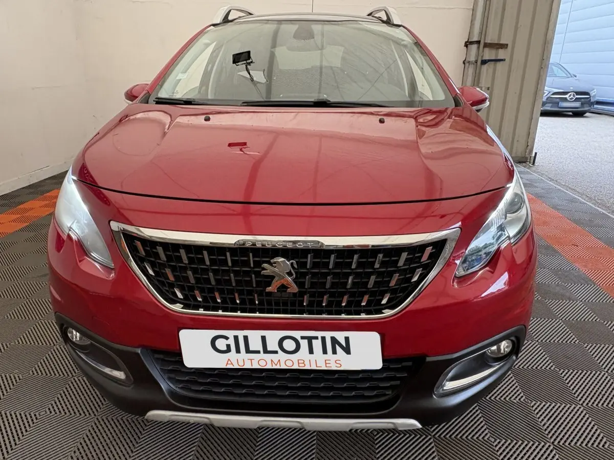 Vue frontale d'un Peugeot 2008 rouge avec calandre chromée et phares allumés dans un garage.