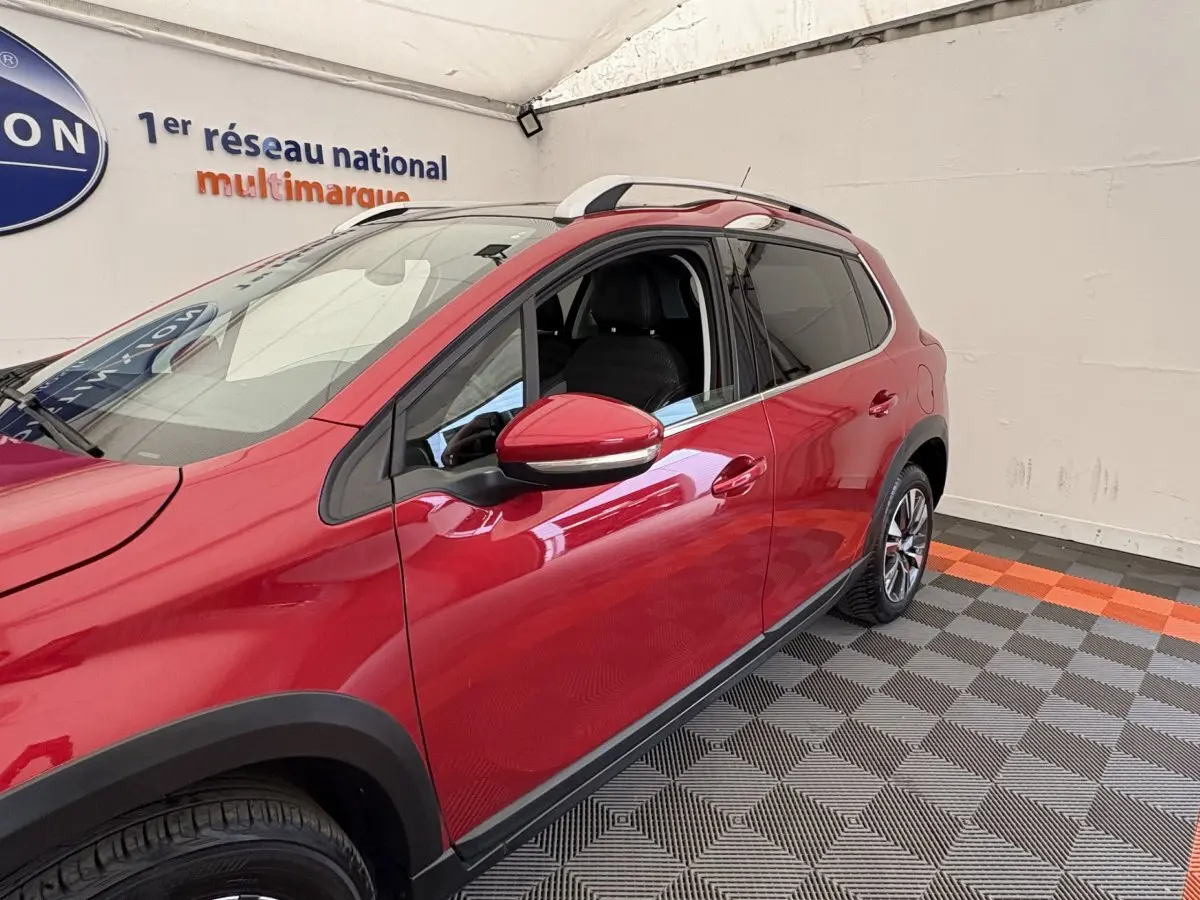 Peugeot 2008 rouge vue de côté gauche en intérieur, avec rétroviseurs chromés et rails de toit visibles.