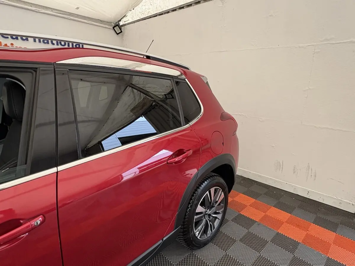 Vue côté arrière droit du Peugeot 2008 rouge avec jantes alliage et barres de toit chromées dans un garage.