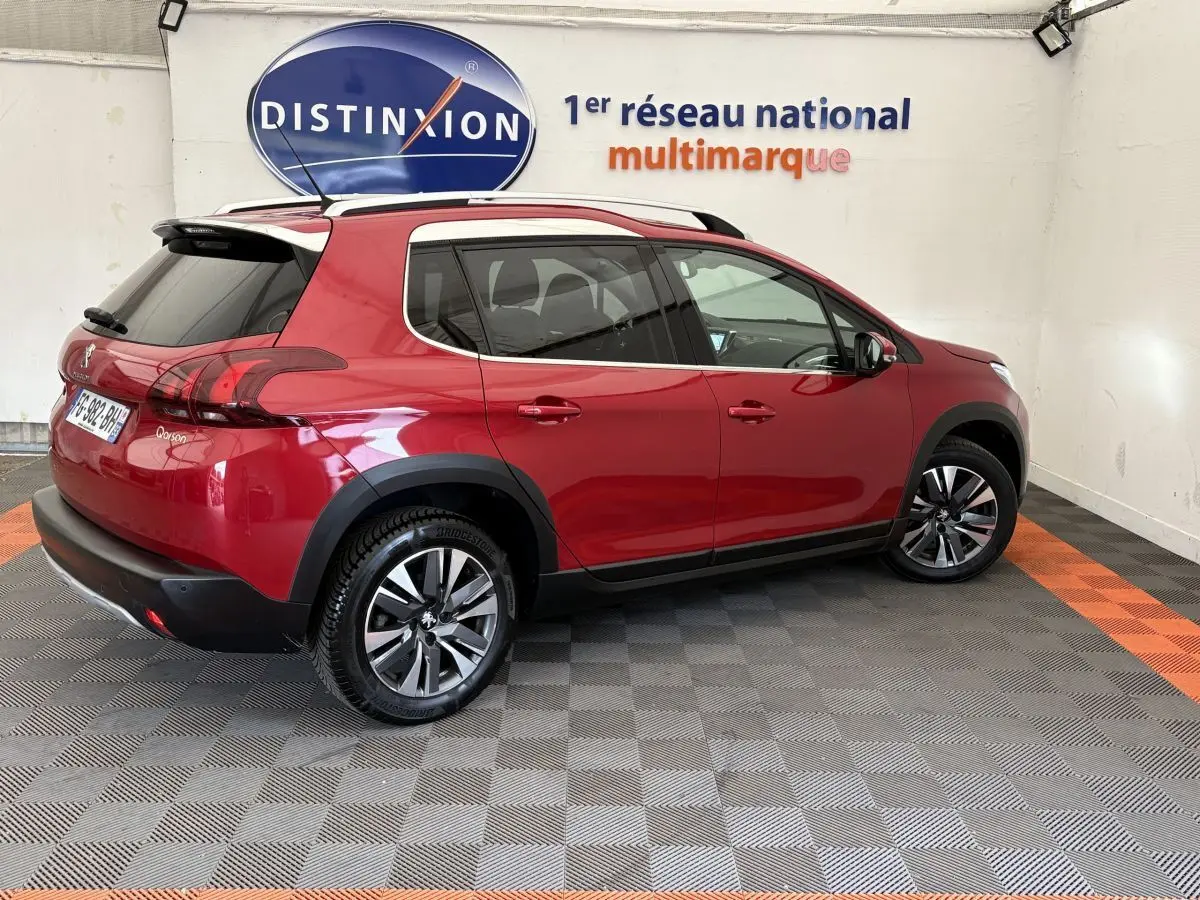 Vue 3/4 arrière droite d'un Peugeot 2008 rouge brillant avec jantes alliage et barres de toit chromées dans un showroom