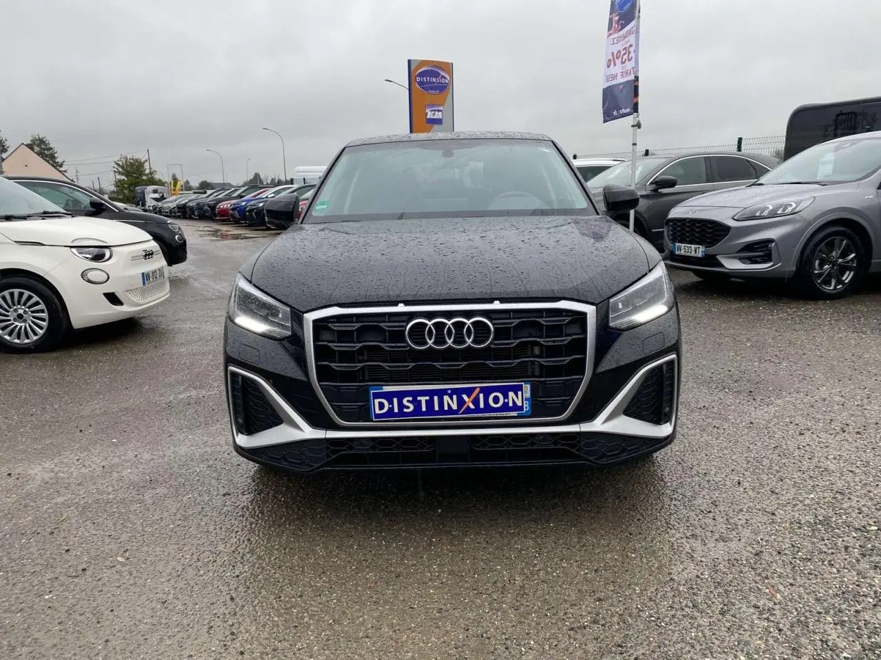 Vue avant d’un Audi Q2 noir 2023 avec calandre large, phares allumés et pluie visible sur la carrosserie.