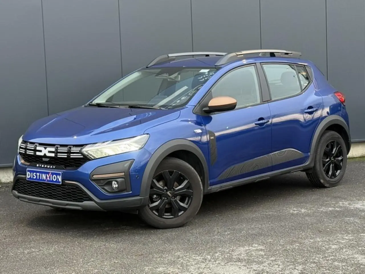 Vue 3/4 avant droite de la Dacia Sandero Stepway Bleu Iron avec jantes noires et barres de toit gris Mégalithe.