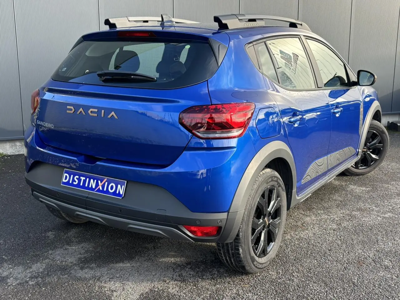 Vue 3/4 arrière droite d'une Dacia Sandero Stepway Bleu Iron avec jantes noires et barres de toit gris Mégalithe.