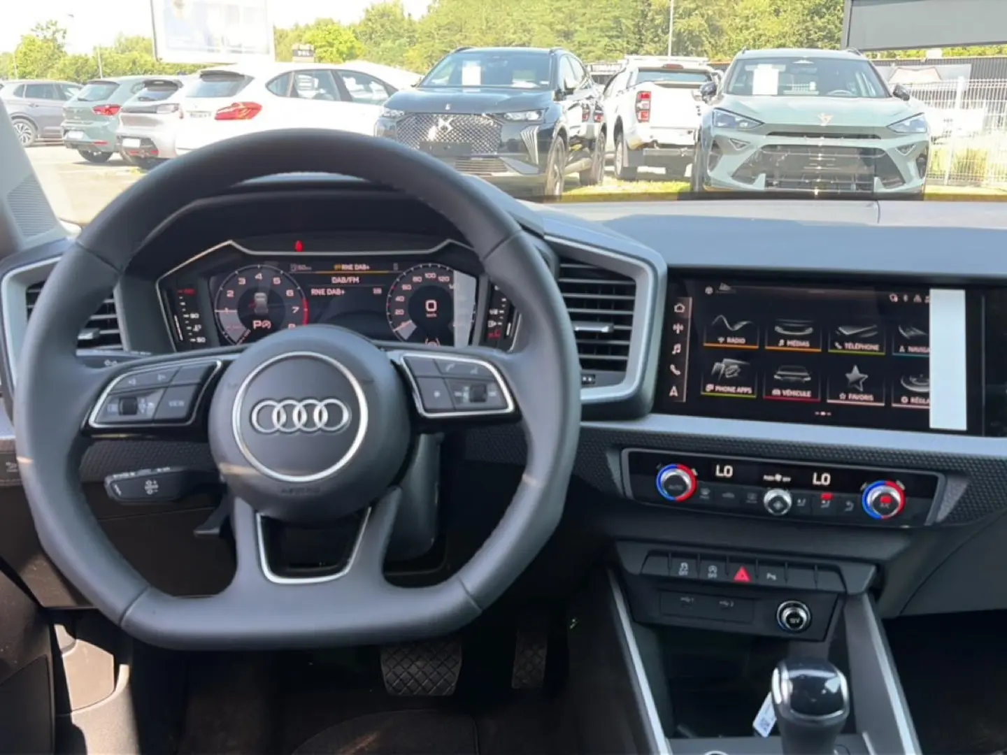 Vue intérieure centrée sur le volant Audi et tableau de bord numérique, avec écran tactile et commandes modernes visibles.