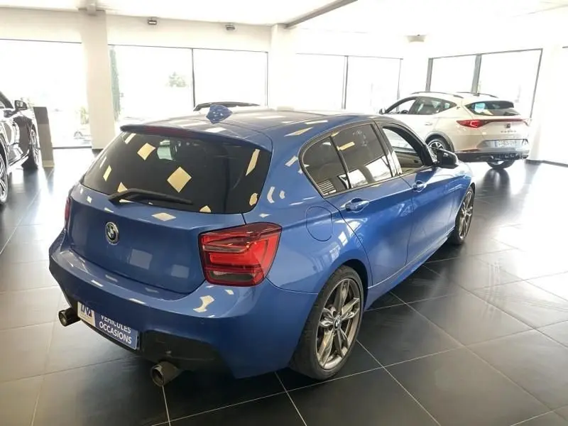 BMW Série 1 135 i bleu métal vue 3/4 arrière droit en intérieur, avec jantes sport et double sortie d’échappement.
