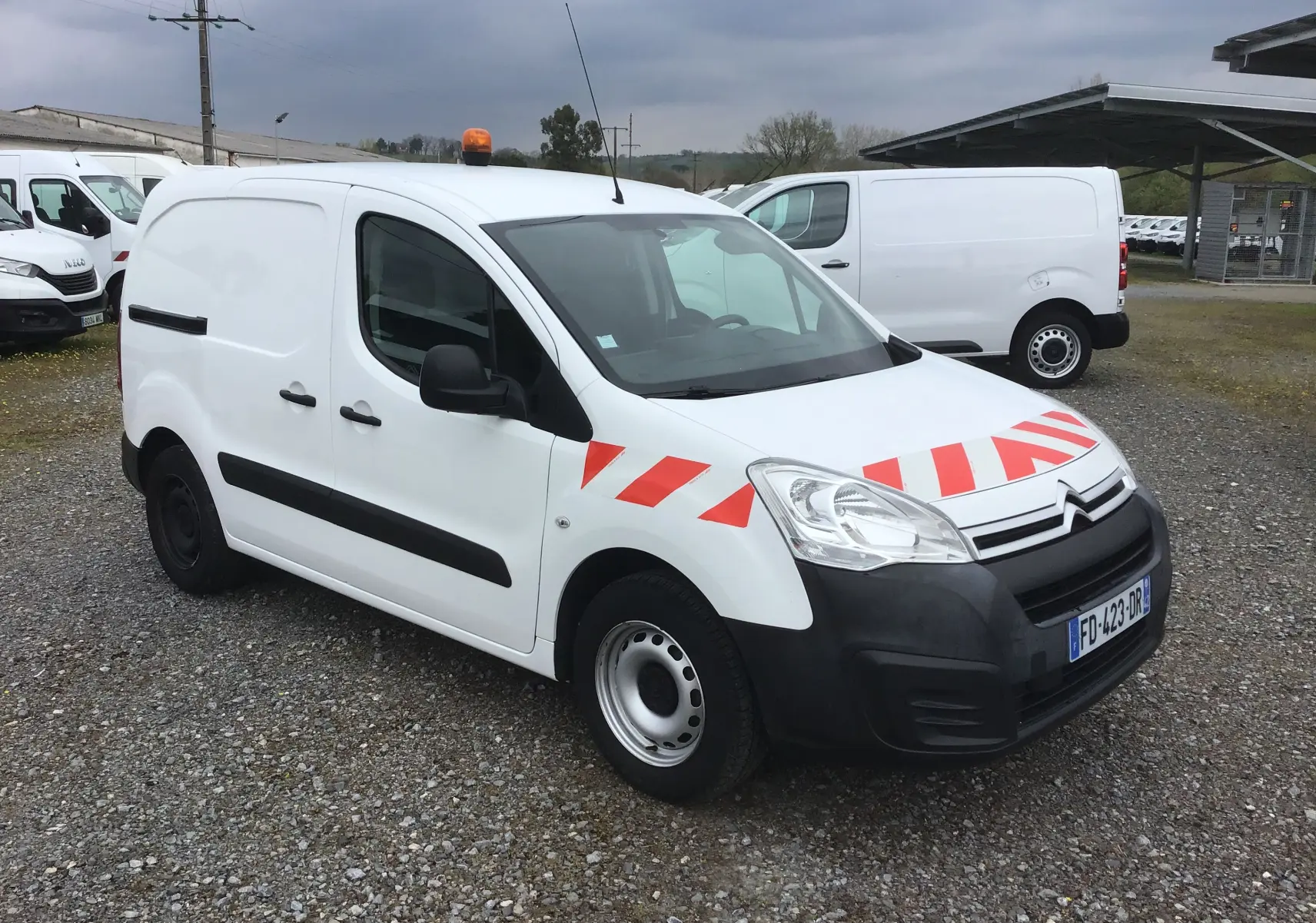 Vue 3/4 avant d’un Citroën Berlingo blanc avec bandes rouges diagonales sur le capot et gyrophare orange sur le toit.