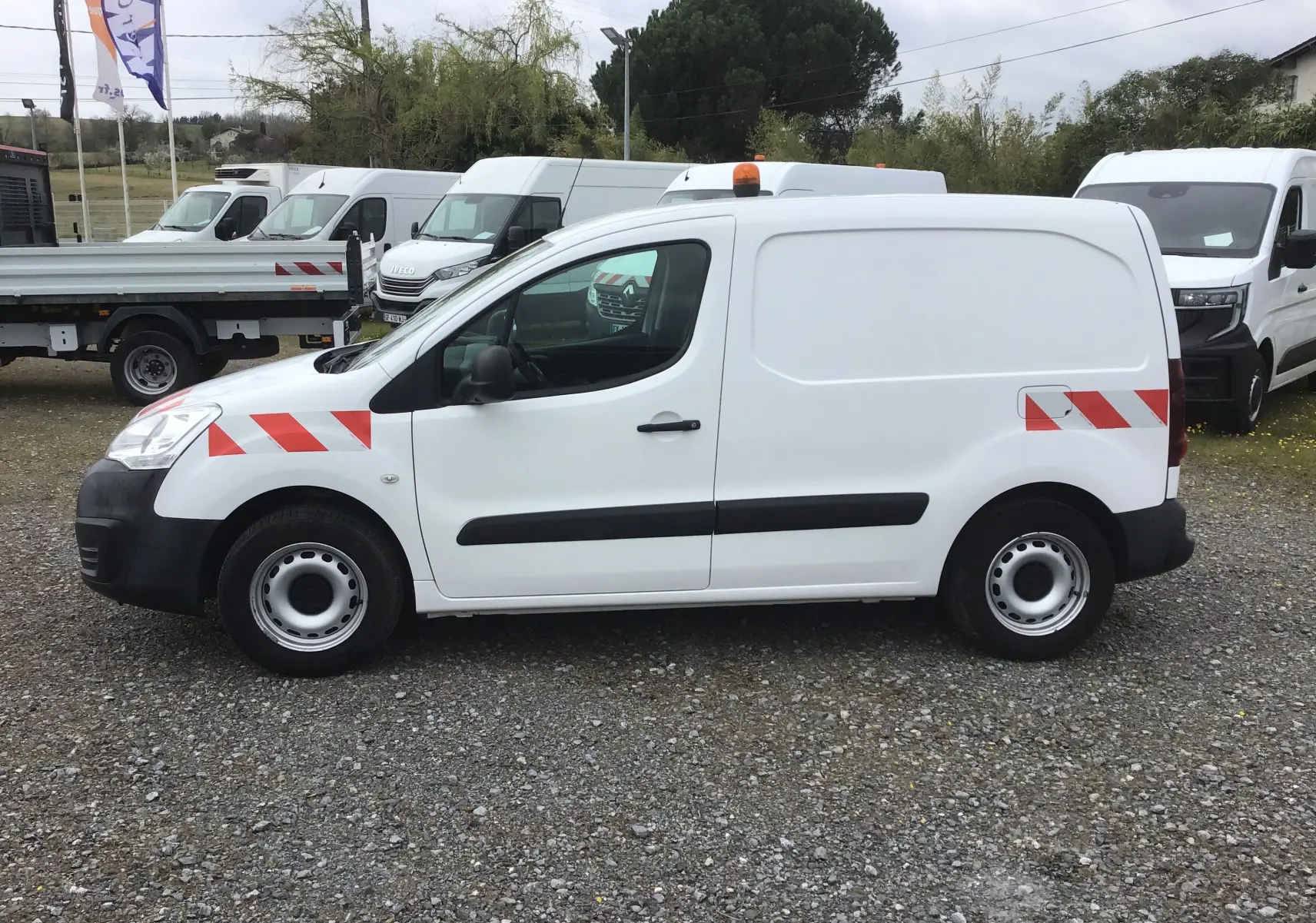 Vue de profil côté gauche d’un utilitaire Citroën Berlingo blanc de 2019 avec bandes rouges réfléchissantes et gyrophare orange.