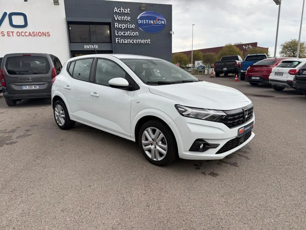 Dacia Sandero blanc vue 3/4 avant droit, garée devant un bâtiment commercial avec plusieurs autres voitures visibles.