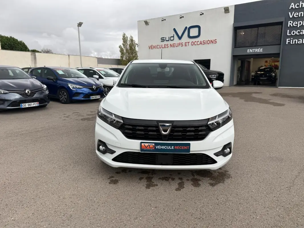Vue frontale d'une Dacia Sandero blanche 2021 avec calandre noire et feux antibrouillard sur un parking de concession.