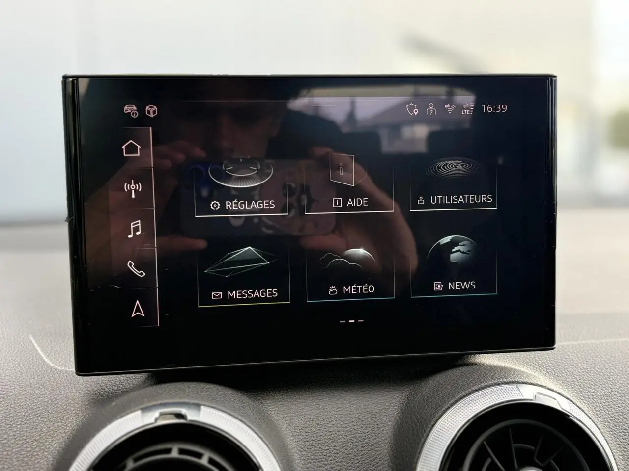 Écran tactile central de l'Audi Q2 35 TDI 150 S-Tronic S-Line affichant le menu principal avec options réglages, aide et météo.