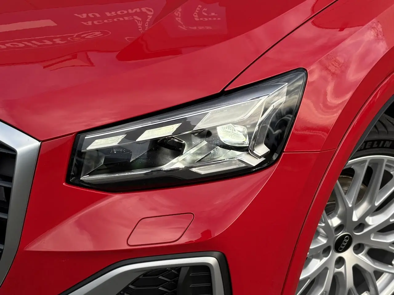Gros plan sur le phare avant droit et la roue argentée d’un Audi Q2 rouge métallisé 2025 finition S-Line.