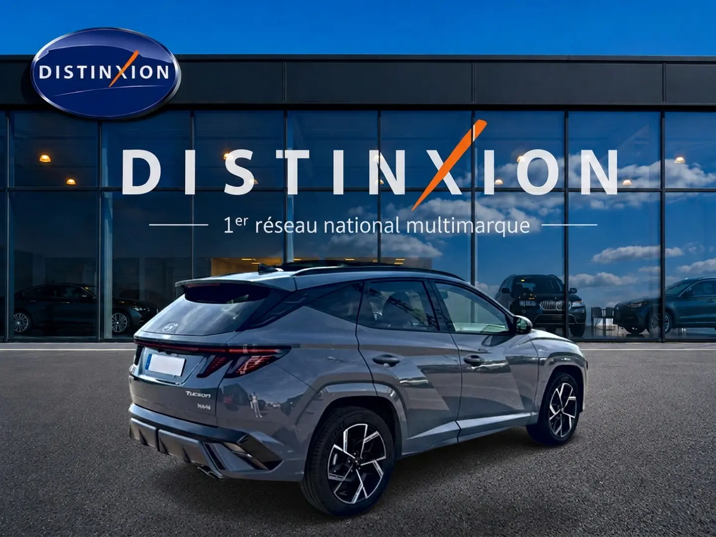 Vue 3/4 arrière droite du Hyundai Tucson gris shadow avec jantes alu noires et toit panoramique devant un showroom Distinxion.