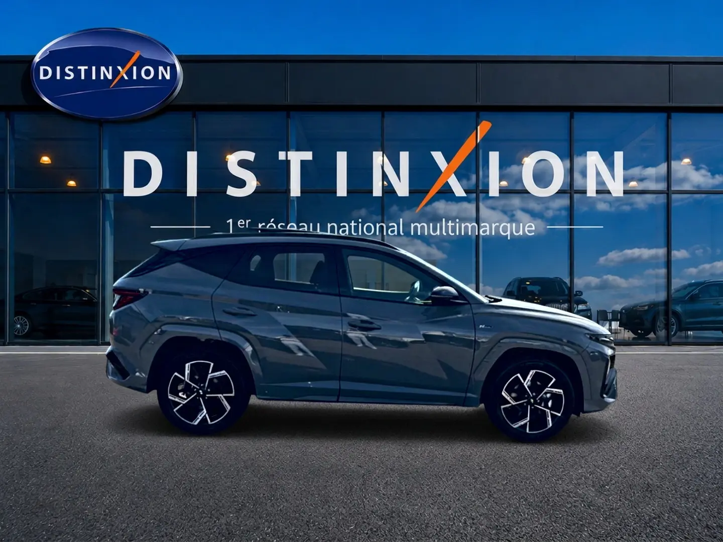 Hyundai Tucson 2026 gris shadow en profil droit devant une vitrine Distinxion, jantes alu noires et chromées visibles.