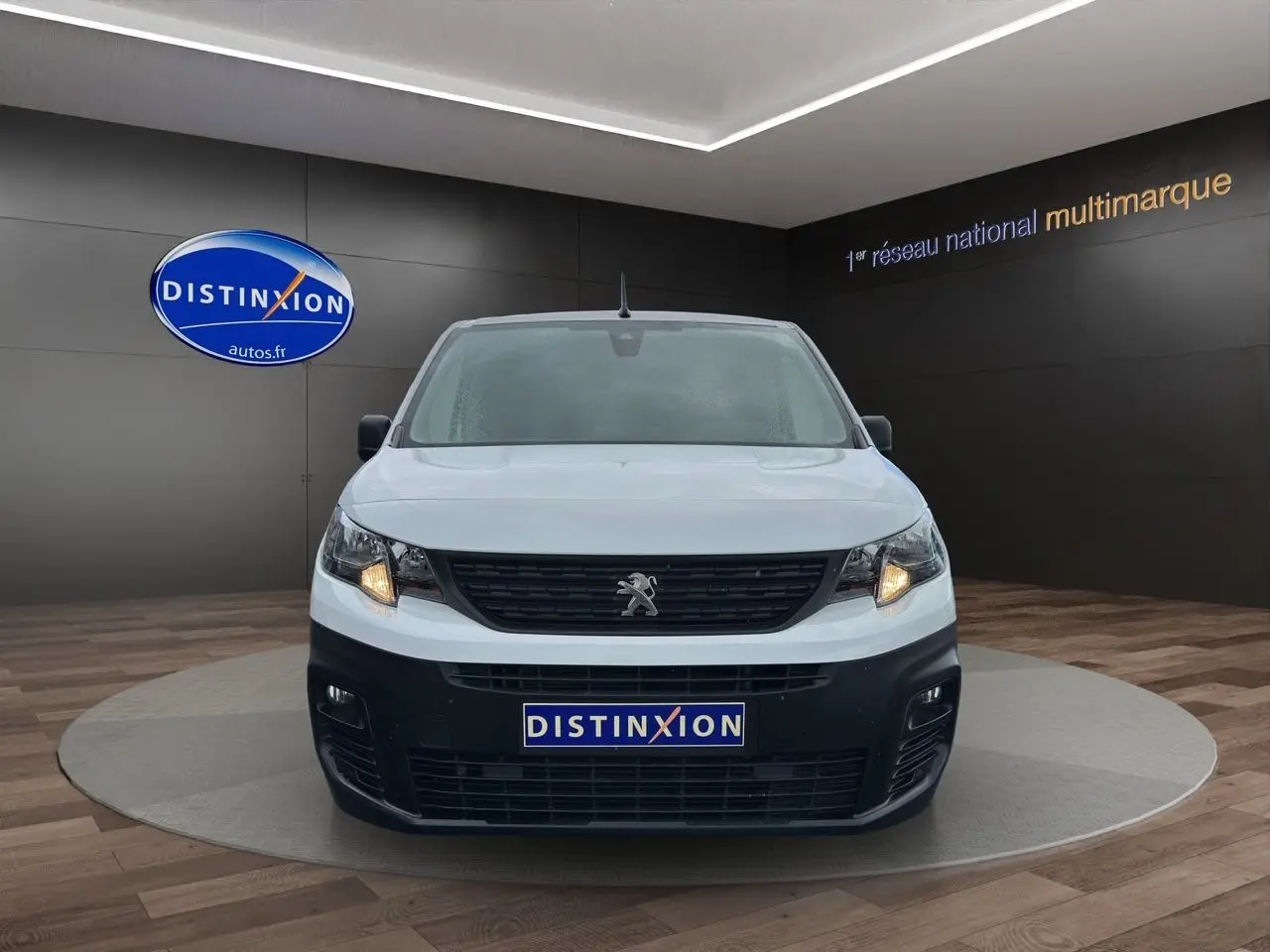 Vue frontale d'un Peugeot Partner blanc 2022 avec calandre noire et phares allumés en showroom.