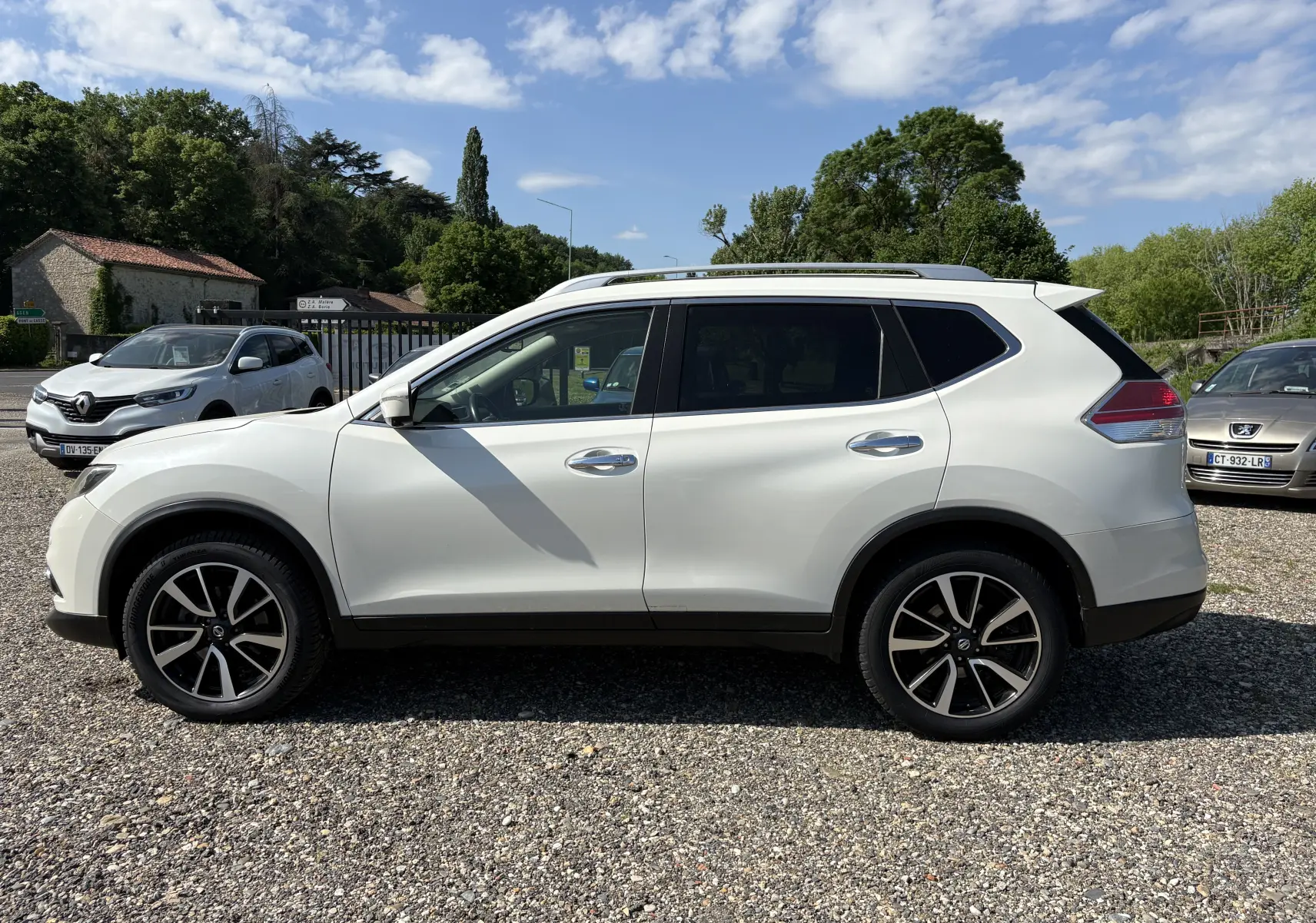 Nissan X-Trail blanc en 3/4 arrière droit, avec jantes noires et toit ouvrant, stationné sur un sol gravillonné.