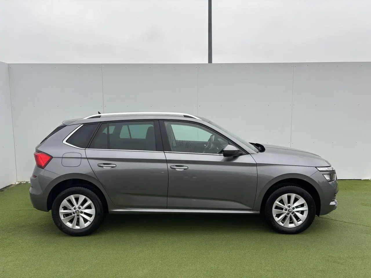 Profil droit d'un Skoda Kamiq gris graphite métallisé avec jantes alliage 17 pouces sur fond vert et mur blanc.