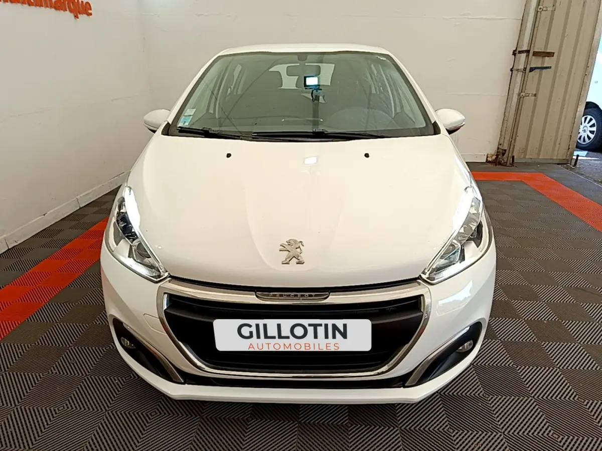 Vue avant d'une Peugeot 208 Puretech 82 Signature blanche stationnée en intérieur avec calandre noire et phares allumés.