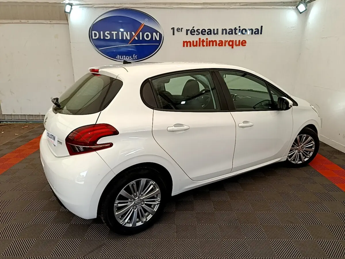 Vue 3/4 arrière droite d'une Peugeot 208 Puretech 82 Signature blanche en intérieur sur sol damier rouge et gris.