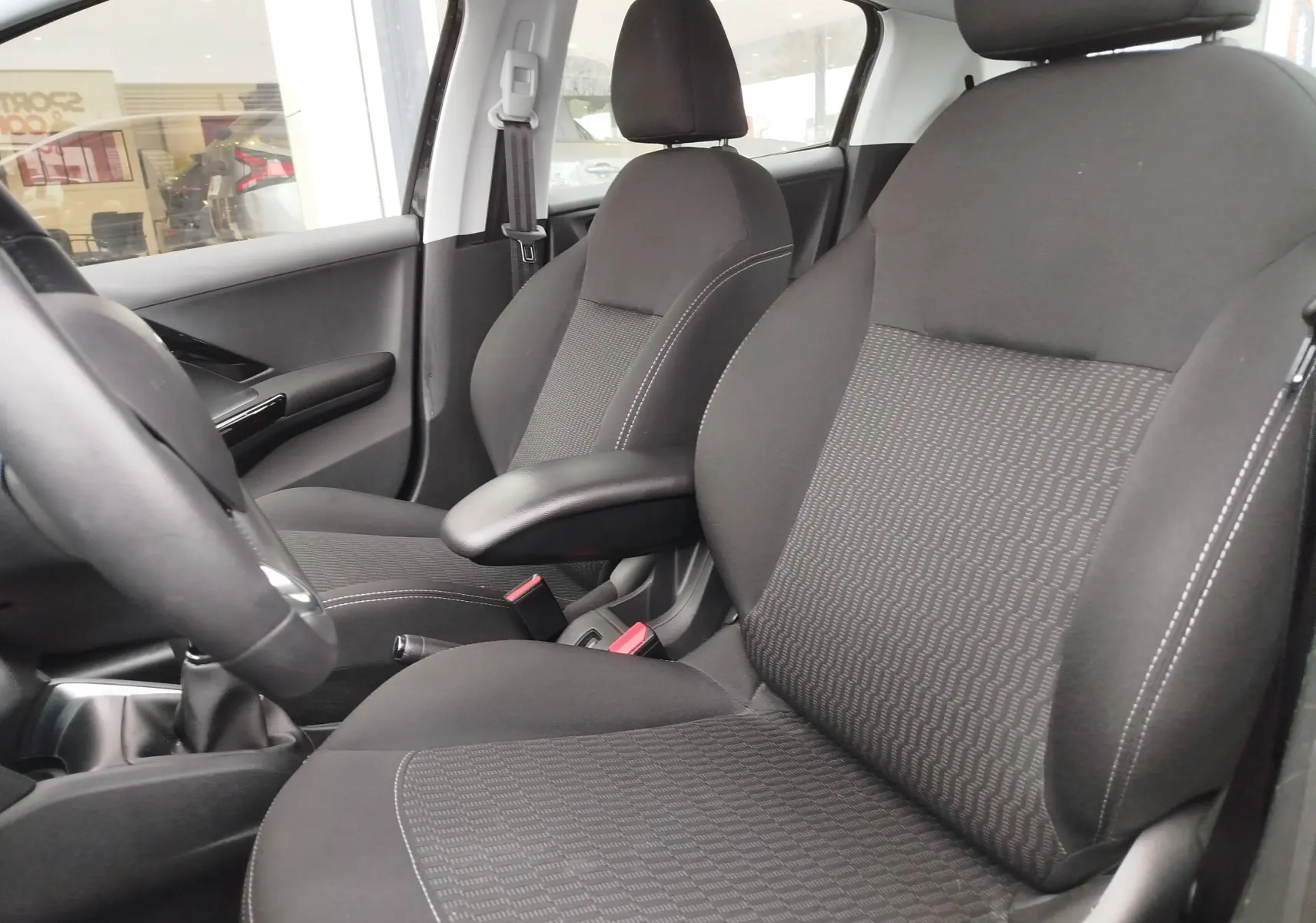 Vue intérieure côté conducteur de la Peugeot 208 2019, sièges noirs avec accoudoir central et volant partiel visible.