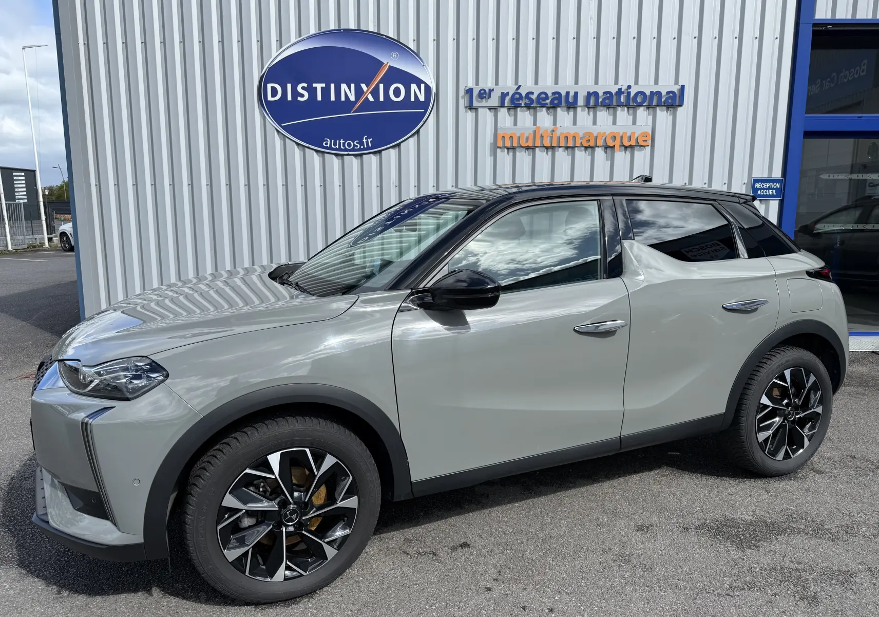 Vue latérale droite d'une DS3 2024 gris laqué avec toit noir, jantes noires et détails modernes devant un bâtiment Distinxion.
