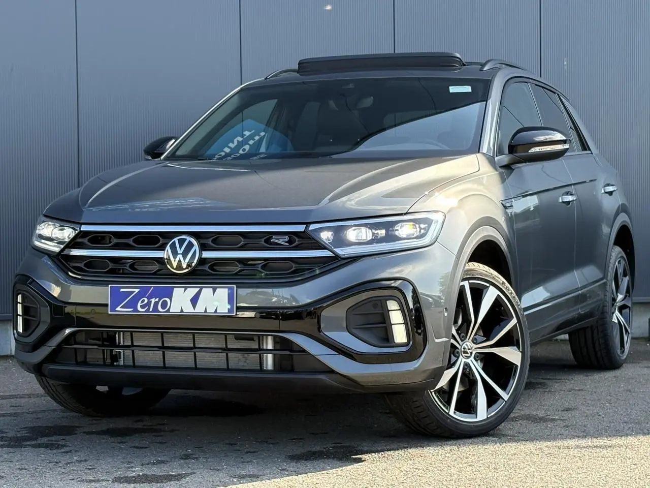 Volkswagen T-Roc gris Indium en 3/4 avant droit avec jantes alliage 19 pouces et toit ouvrant visible.