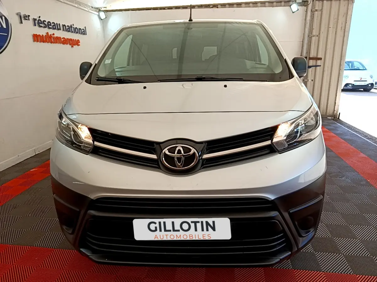 Vue frontale d'un Toyota Proace Verso gris clair avec phares allumés et calandre noire dans un showroom.
