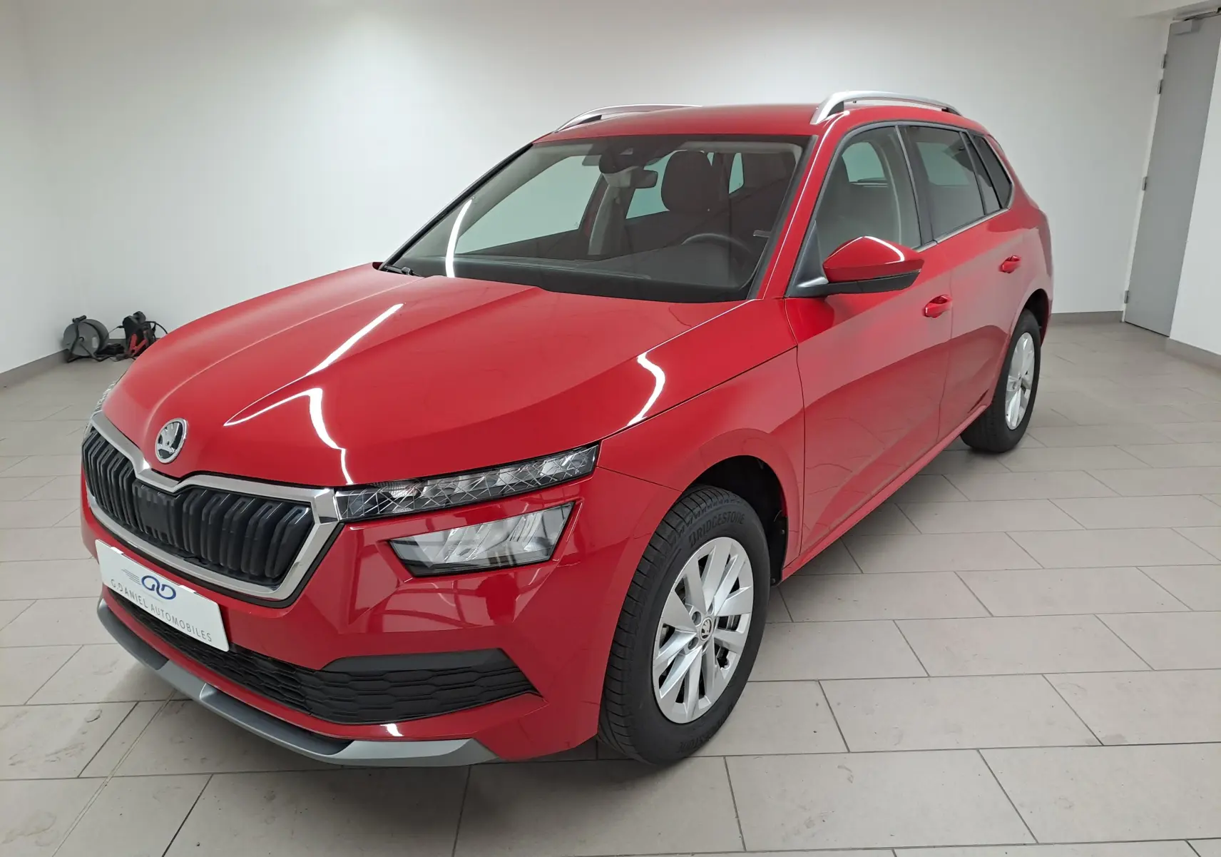 Vue 3/4 avant droite d'un Skoda Kamiq 2024 rouge Velvet avec calandre noire et jantes argentées en intérieur showroom.