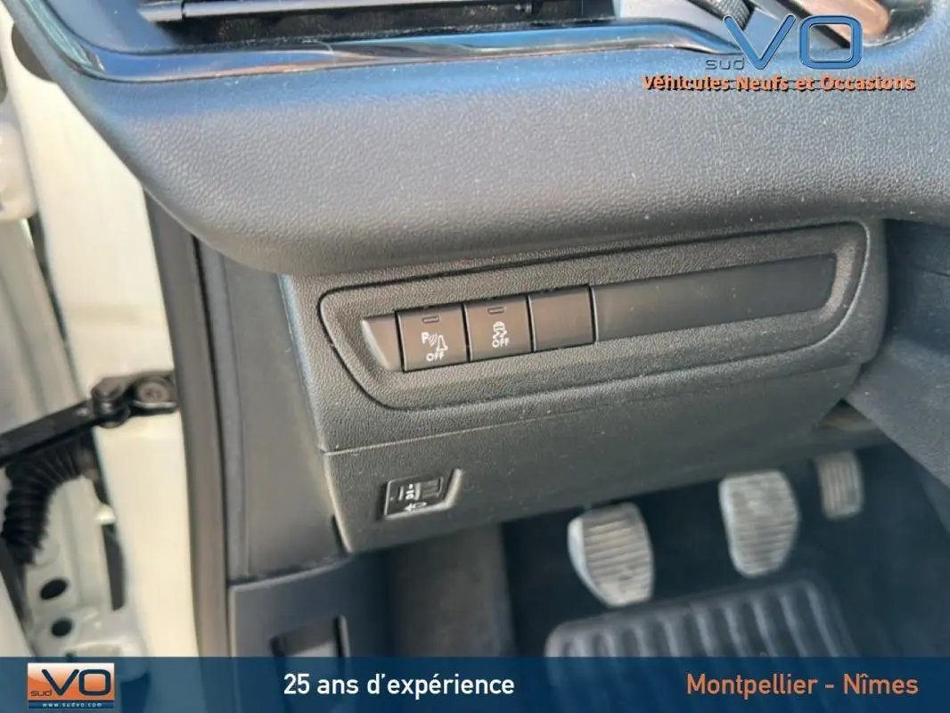Détail des commandes sous le tableau de bord côté gauche d'une Peugeot 208 blanche de 2015 avec pédales et portière ouverte.