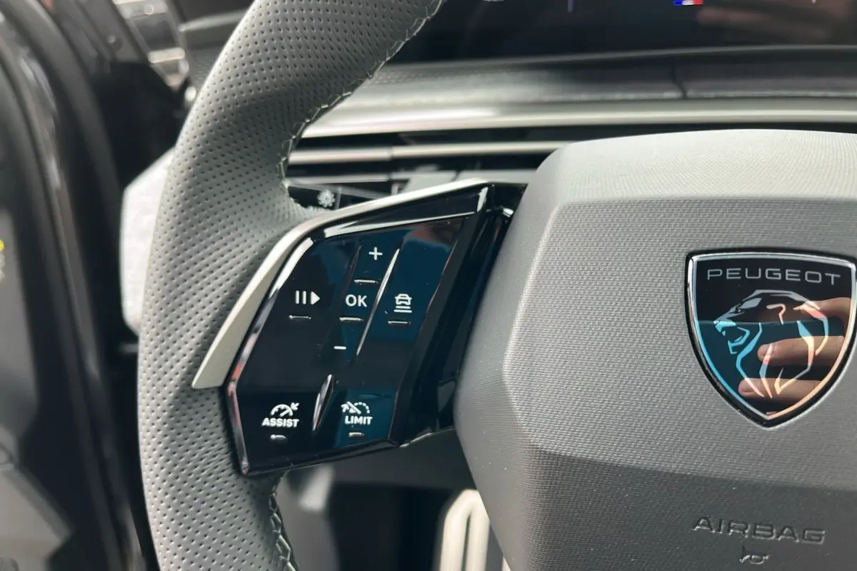 Gros plan sur le volant cuir perforé côté gauche du Peugeot 5008 Hybrid 145 avec logo Peugeot et commandes intégrées.