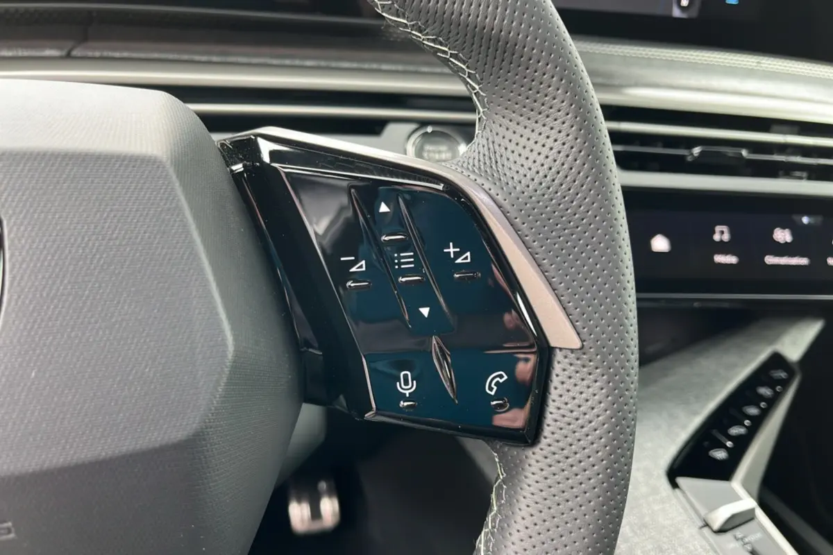 Gros plan sur les commandes gauche du volant cuir perforé du Peugeot 5008 Hybrid 145 gris titane avec tableau de bord noir.