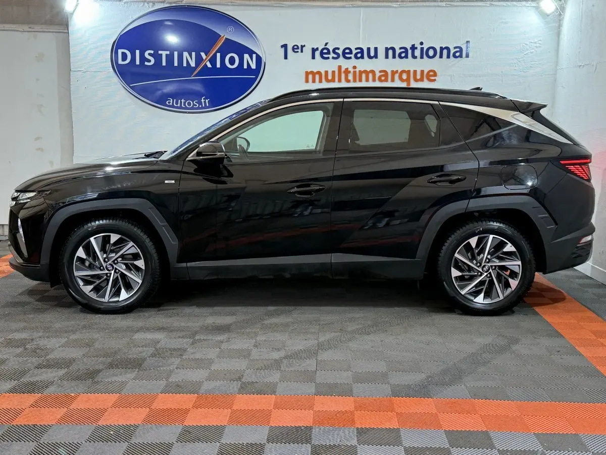 Profil côté gauche d'un Hyundai Tucson noir 2021 hybride, avec jantes alliage et feux arrière LED visibles en intérieur showroom.