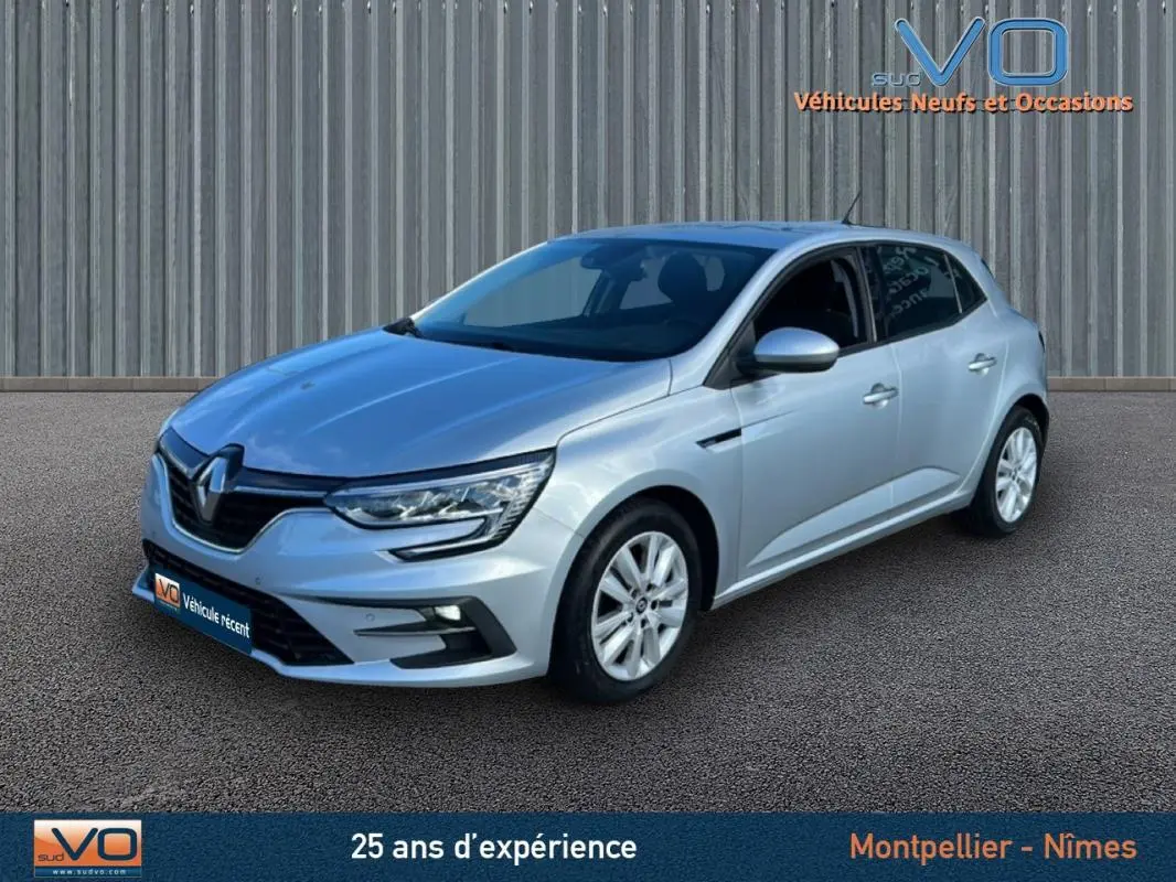 Renault Mégane IV Berline gris clair vue 3/4 avant droit sur sol bitumé devant un mur métallique