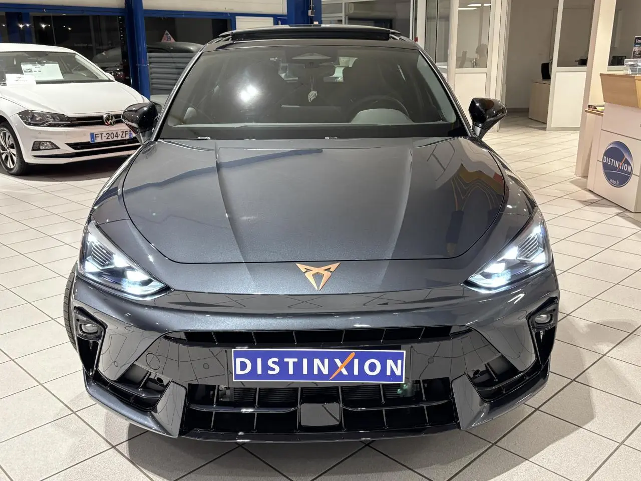 Vue avant d'une CUPRA Leon gris Graphène avec logo cuivré et phares LED allumés en showroom.