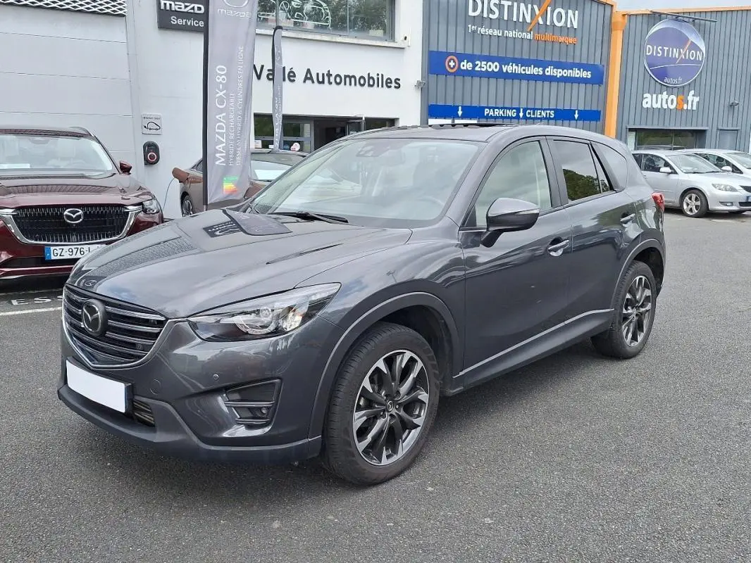 Vue 3/4 avant droit d'un Mazda CX-5 gris métallisé garé devant un concessionnaire avec jantes aluminium noires.