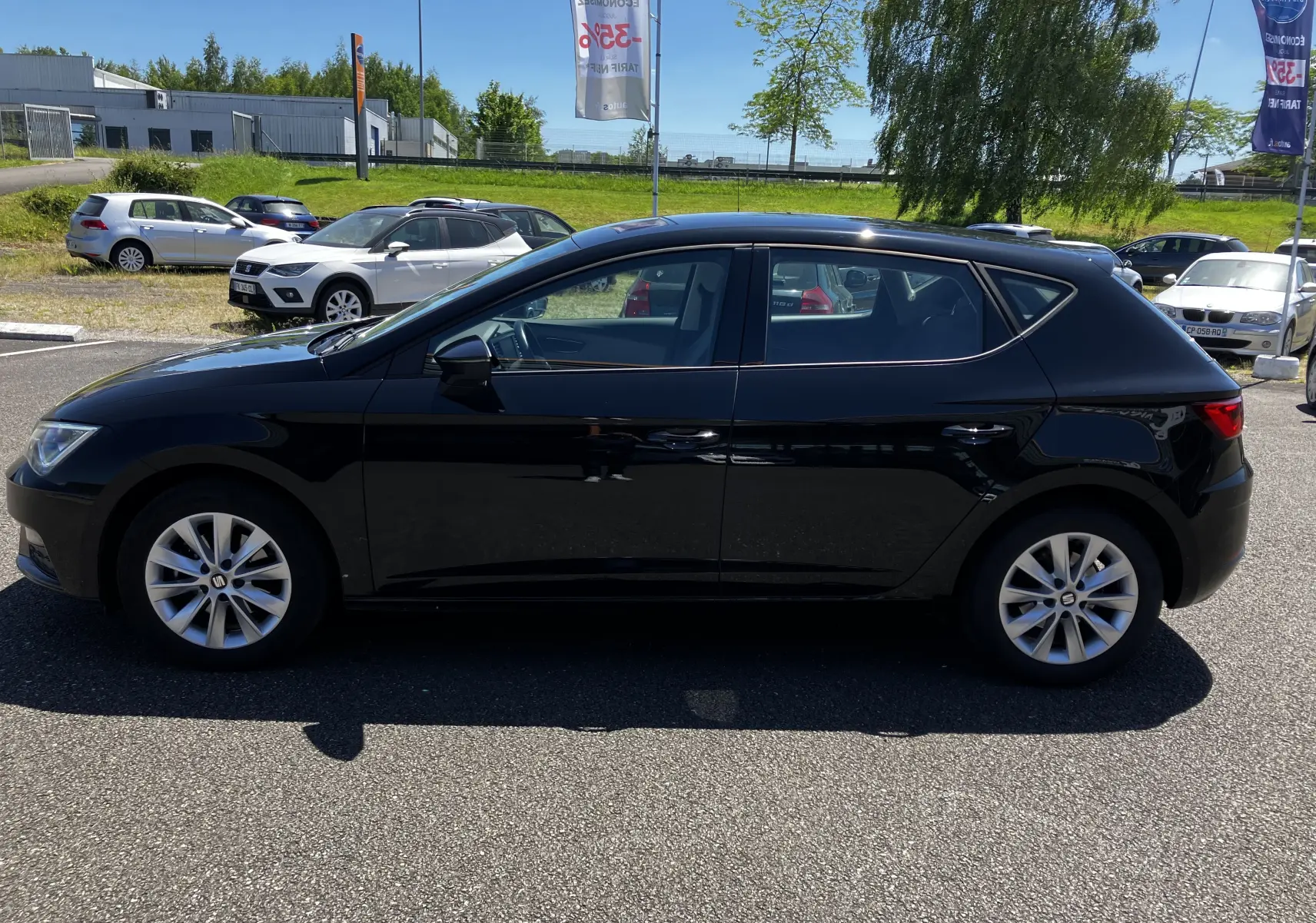 Profil latéral droit d'une SEAT LEON noire 5 portes, garée en extérieur par temps ensoleillé.