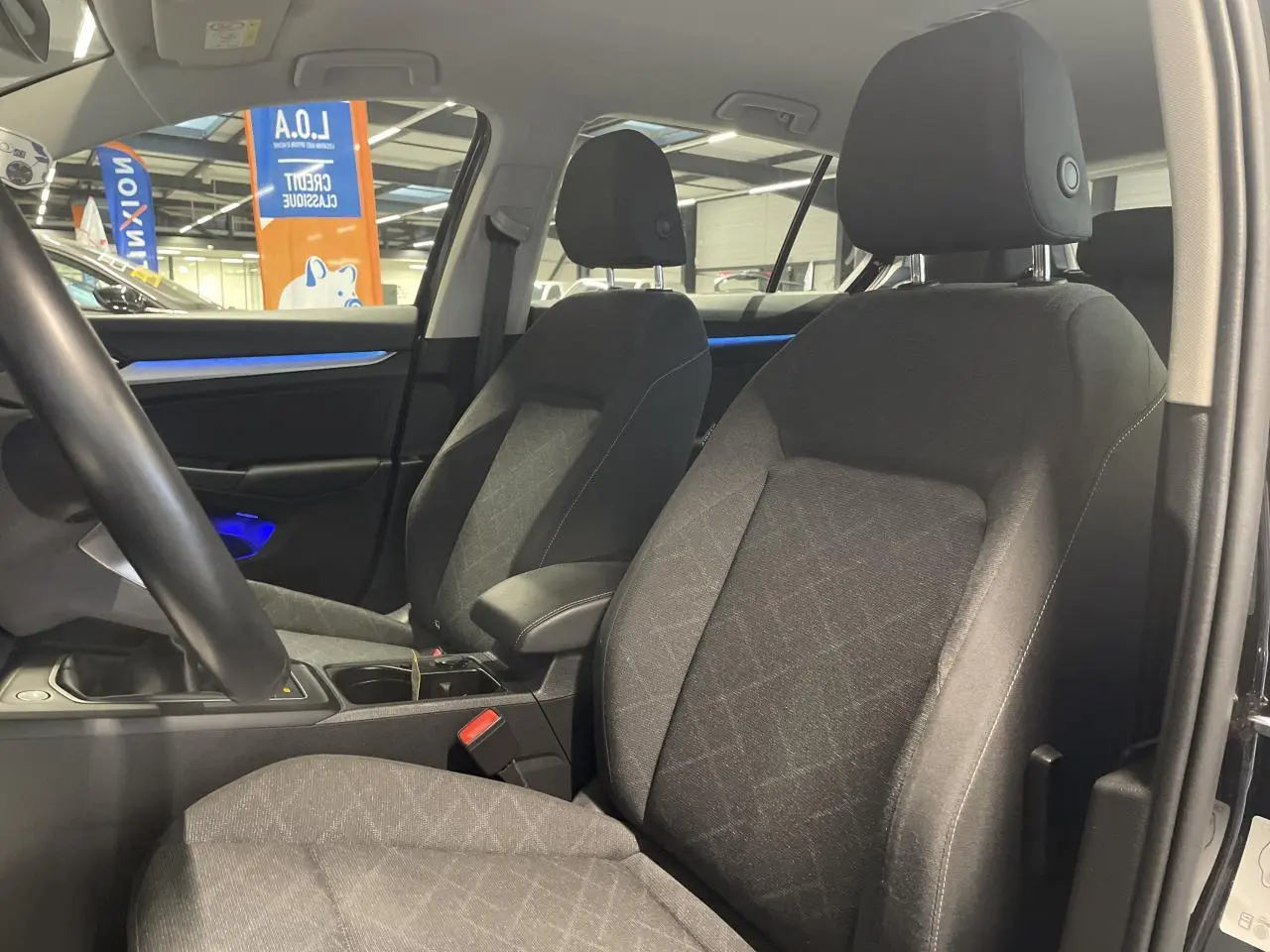 Intérieur avant de la Volkswagen Golf 2024 noir, sièges en tissu gris avec accoudoir central et éclairage d'ambiance bleu.