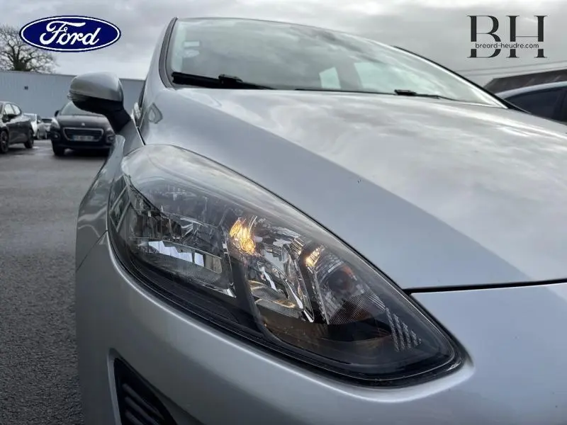 Gros plan sur le phare avant gauche d'une Ford Fiesta gris lunaire, avec rétroviseur et capot visibles.