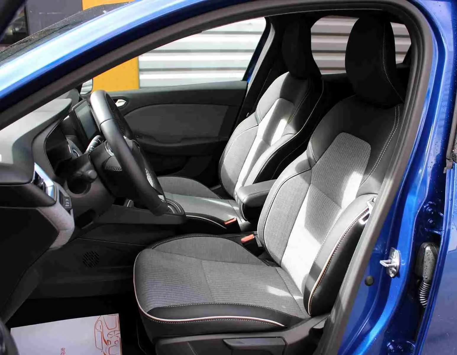 Vue intérieure côté conducteur de la Renault Clio bleu iron metal, volant multifonction et écran tactile central visible.