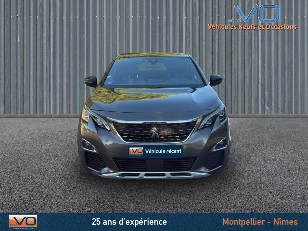 Vue frontale d'un Peugeot 3008 gris foncé avec calandre chromée et phares LED, stationné devant un mur métallique.