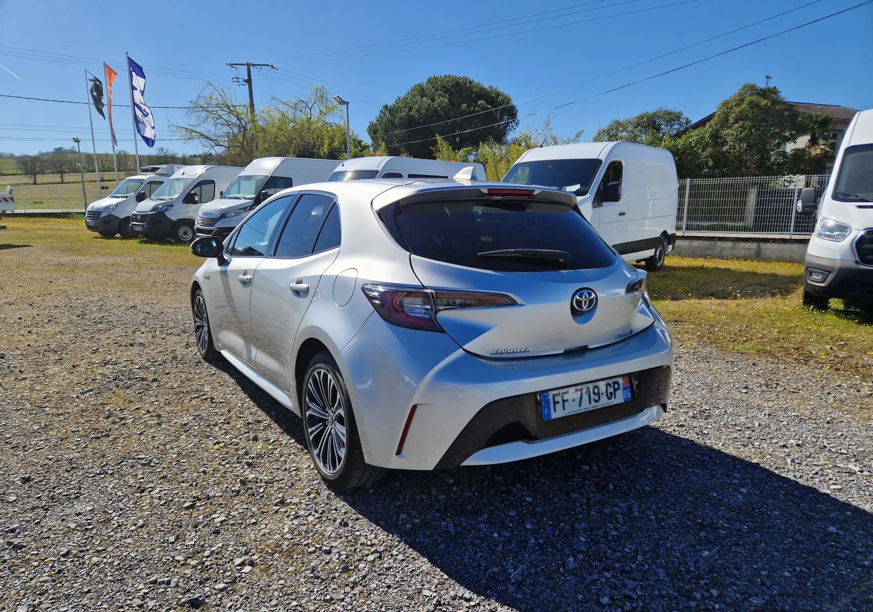 Toyota Corolla hybride gris clair métal vue 3/4 arrière droit sur parking avec plusieurs fourgons blancs en arrière-plan.