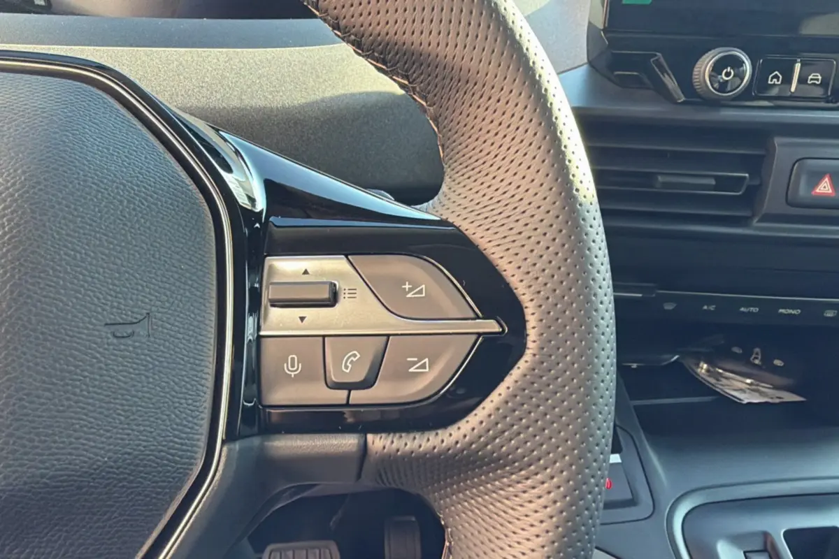 Gros plan sur le volant cuir perforé du Peugeot Rifter 2025, avec commandes multifonctions et tableau de bord noir.