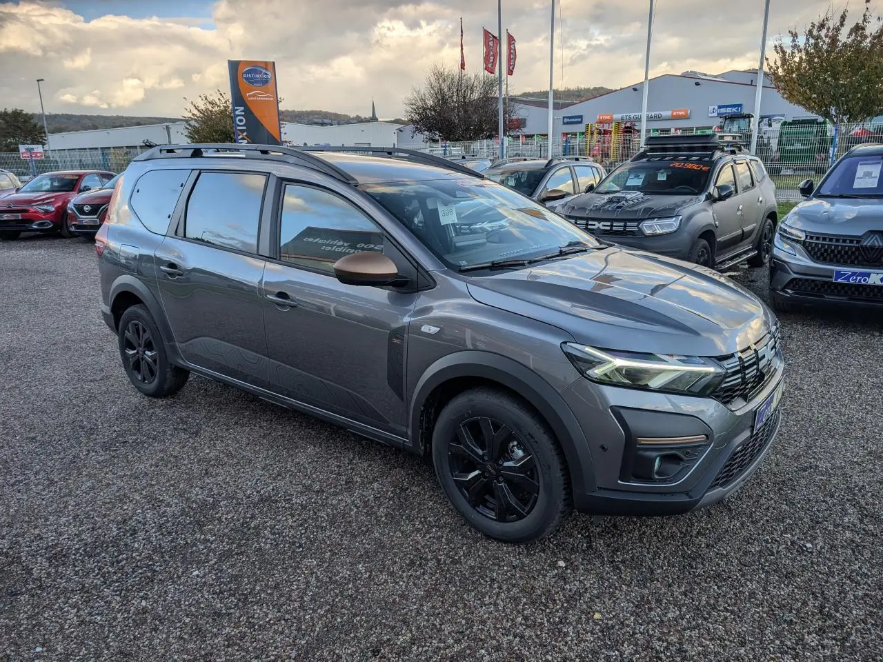 Dacia Jogger gris métallisé en 3/4 avant droit, avec jantes noires et rétroviseurs brun cuivré, stationné en extérieur.