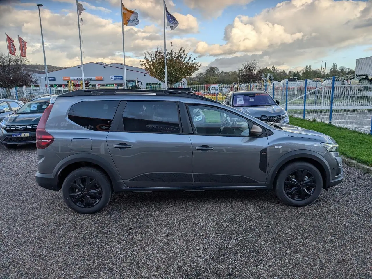 Vue de profil côté gauche d'un Dacia Jogger gris métallisé avec jantes noires et barres de toit sur parking extérieur.