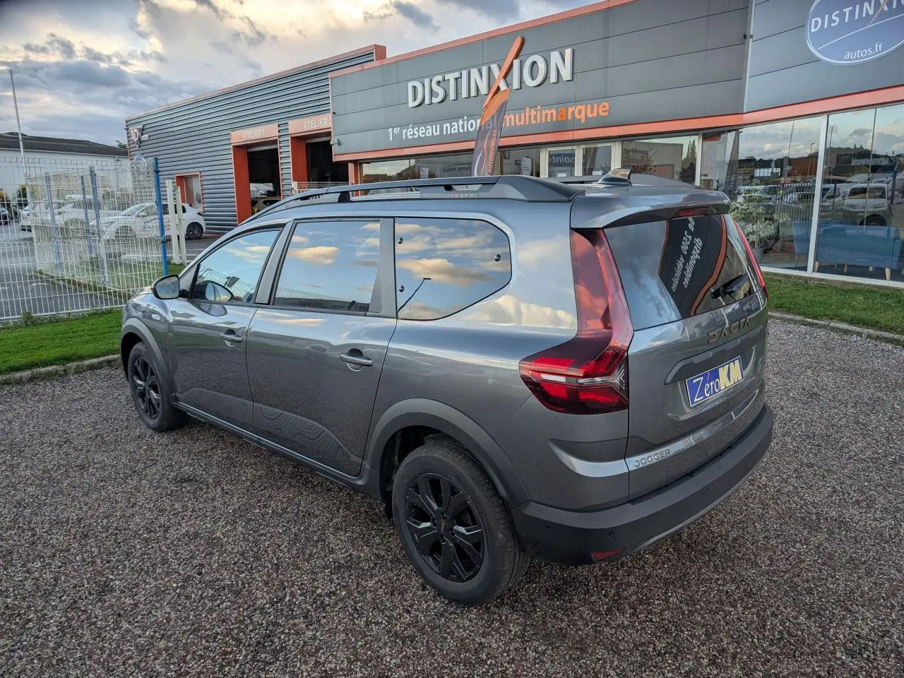 Vue 3/4 arrière droite d'un Dacia Jogger gris métallisé avec jantes noires et barres de toit, stationné devant un concessionnaire.