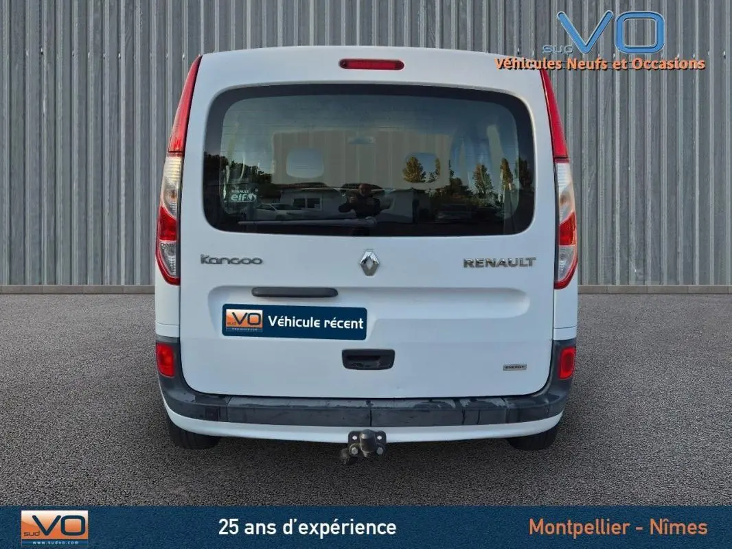 Vue arrière d’un Renault Kangoo blanc 2016 avec attelage et vitres surteintées, garé devant un mur métallique.