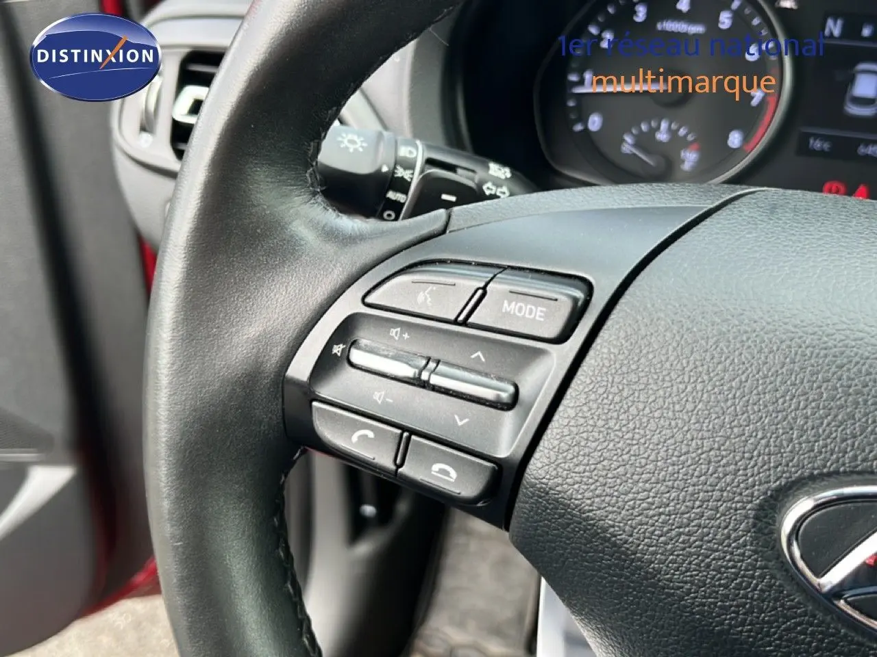 Gros plan sur le volant côté gauche de la Hyundai i30 SW 2023 rouge, avec commandes audio et tableau de bord en arrière-plan.