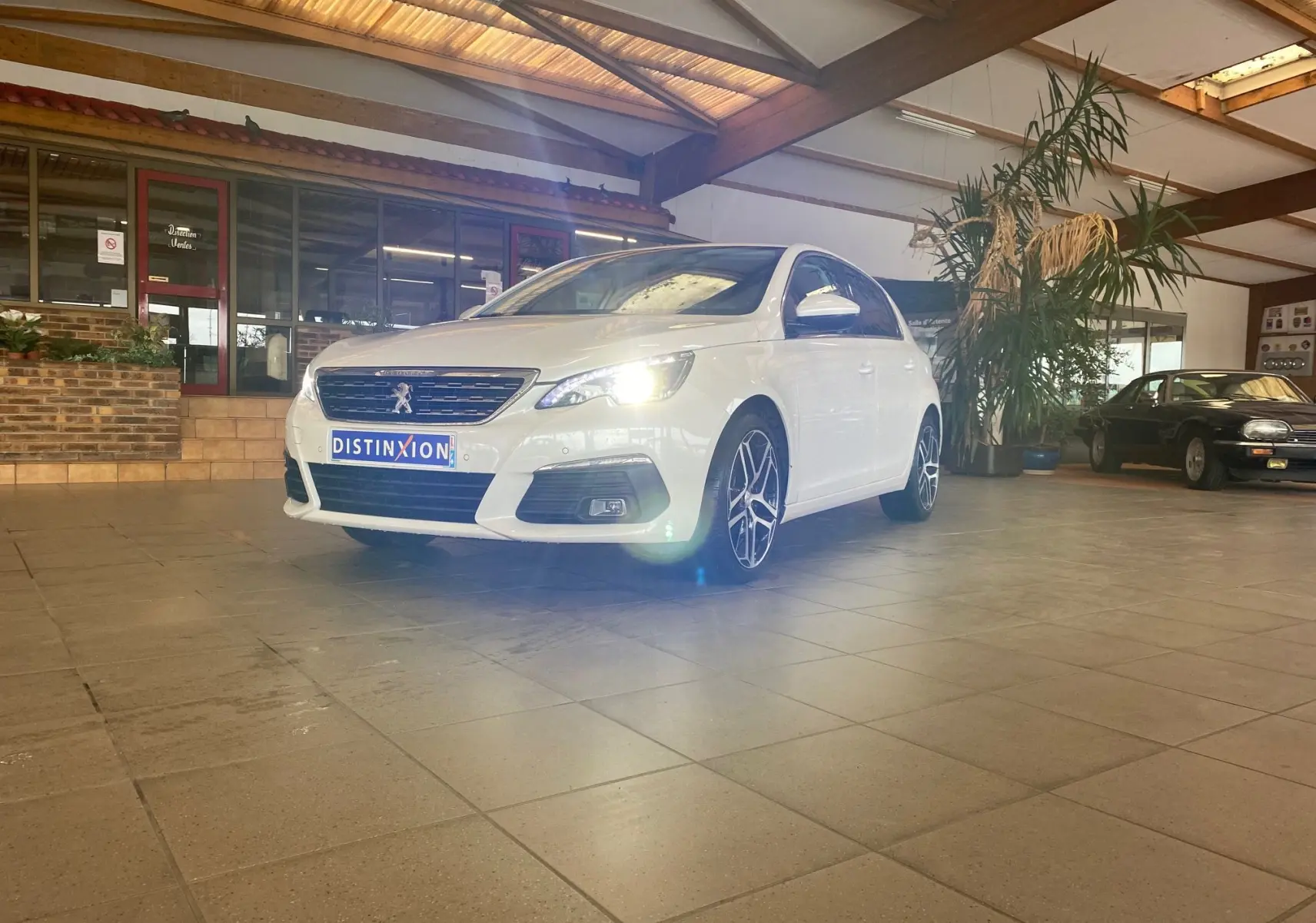 Peugeot 308 blanche vue en 3/4 avant droit dans un showroom, phare avant droit allumé et jantes alliage visibles.