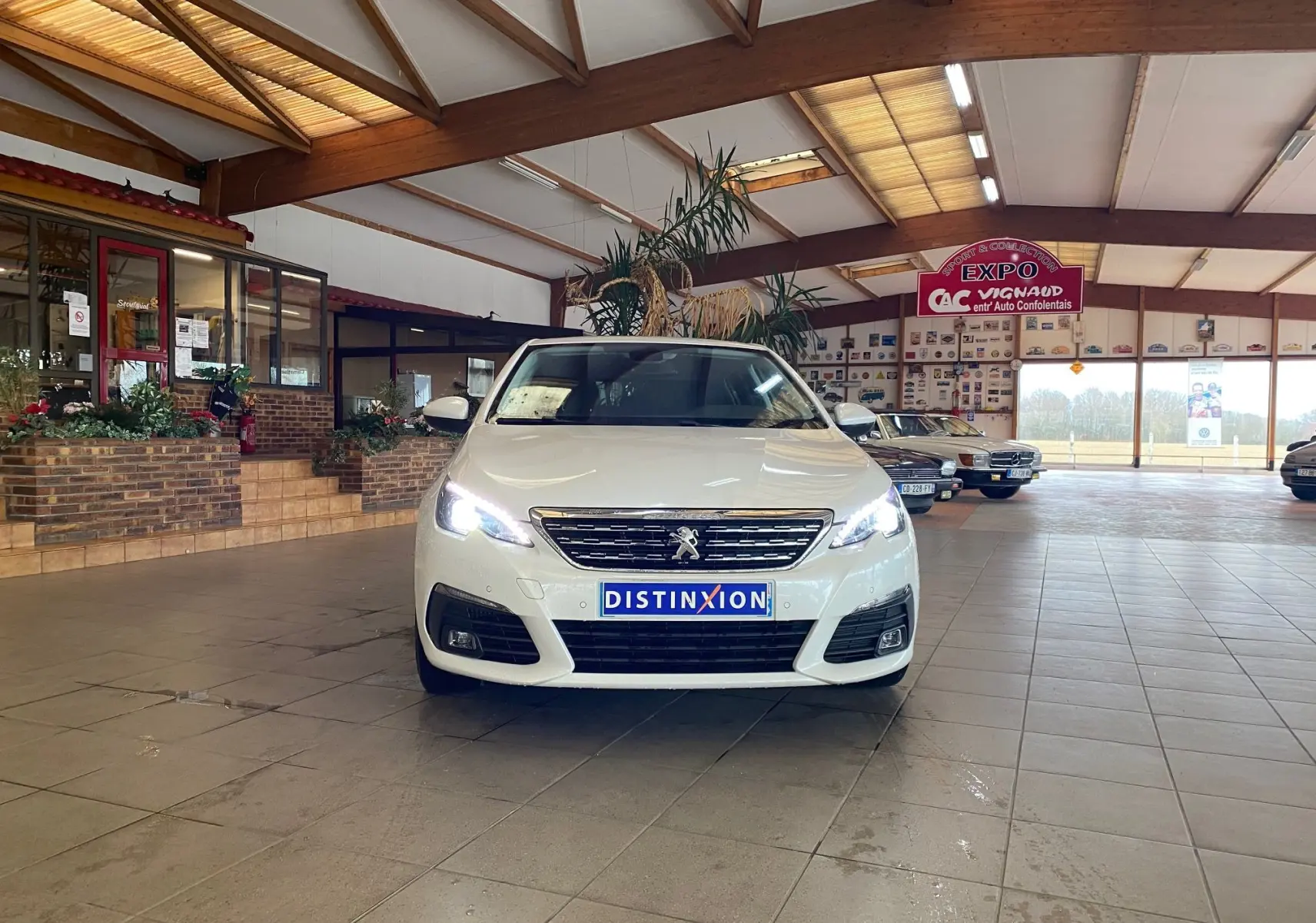 Peugeot 308 blanche vue de face en intérieur avec phares allumés et plaque Distinxion visible.